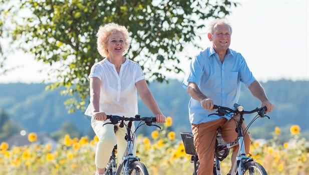 Illustrasjonsfoto av en mann og kvinne som sykler på en sti med en blomstereng og trær i bakgrunnen.