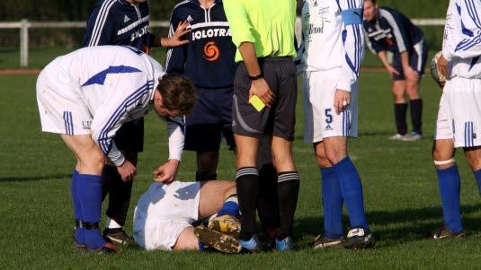 fotballskade, idrettsskade En fotballspiller har fått noe ut av ledd og ligger på banen, mens dommer og medspillere står rundt.