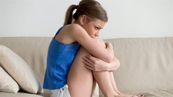 Skam Illustrasjonsfoto av en kvinne som sitter i fosterstilling i sofaen. Kvinnen ser bekymret ut.