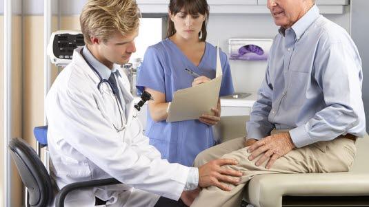 Kneundersøkelse hos lege Fotoet viser en yngre mannlig lege som undersøker det venstre kneet til en eldre mann. Mannen sitter på en undersøkelsesbenk ikledd skjorte og bukse. Imellom og bak legen og pasienten står en helsesekretær, som skriver notater.