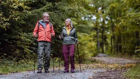 eldre på tur På en skogsti går et par i pensjonsalderen, utstyrt med turlær og staver. De ser ut til å ha det fint mens de går tur og nyter naturen rundt seg.