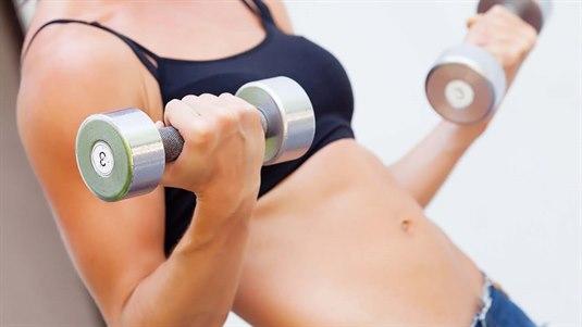 Trening En kvinne løfter vekter på treningsstudioet. Hun holder en manual i hver hånd og utfører Bicepscurls med manualer. Hun har god kroppsholdning.