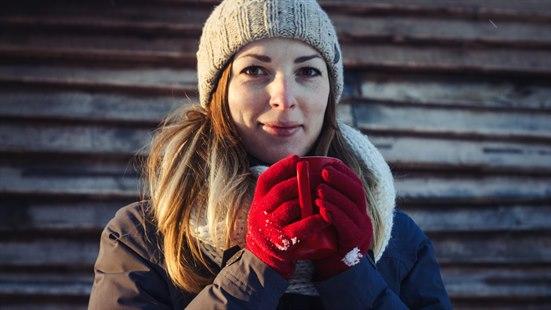 Kalde hender Illustrasjonsfoto: Kvinne med lue, røde votter og brun vinterjakke holder en rød kopp. Hun står foran en tømmervegg og ser inn i kamera.