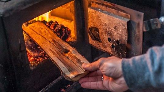 peis En person legger en vedkubbe inn i et fyringsanlegg. Flammene i ovnen skinner på kubben og sender en varm glød.