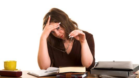 Hodepine En dame sitter ved sin kontorpult og holder brillene sine i den ene hånden mens hun holder seg for pannen